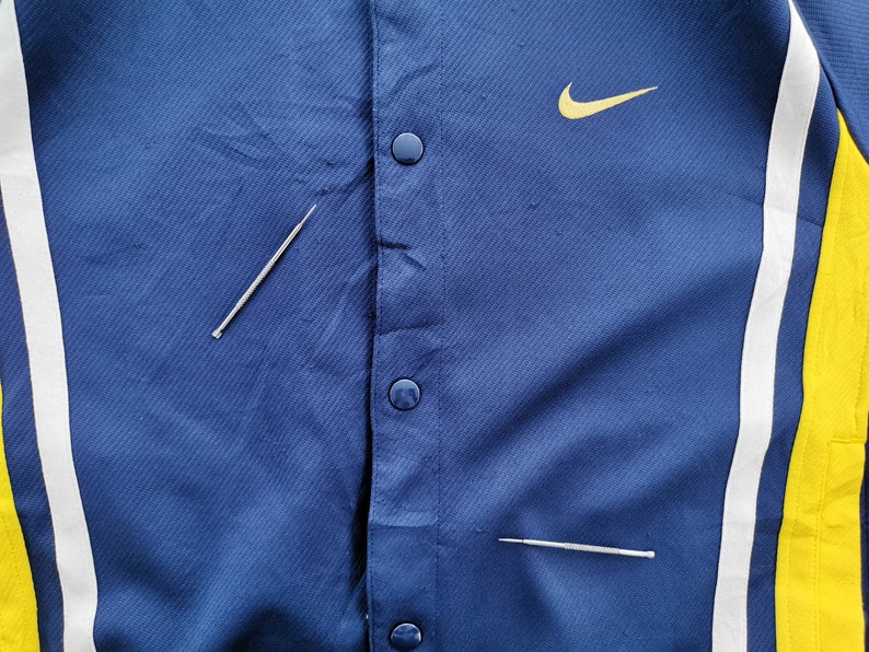 May include: A close-up of a blue jacket with a yellow Nike logo and snap closures. The jacket has white and yellow stripes on the sides. Two silver pins are placed on the jacket.