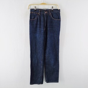 Op de afbeelding: Donkerblauwe spijkerbroek hangend aan een houten hanger. De jeans heeft een klassiek ontwerp met vijf zakken en koperkleurige klinknagels en stiksels. De rechte pijpen zijn zichtbaar en de jeans lijken ongedragen.