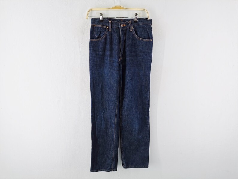 Op de afbeelding: Donkerblauwe spijkerbroek hangend aan een houten hanger. De jeans heeft een klassiek ontwerp met vijf zakken en koperkleurige klinknagels en stiksels. De rechte pijpen zijn zichtbaar en de jeans lijken ongedragen.