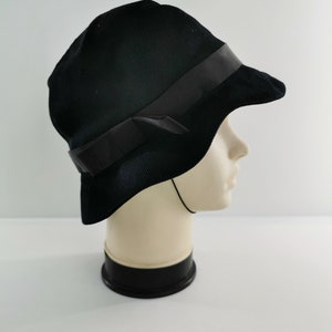 May include: A black cloche hat with a dark ribbon band and bow detail. The hat is made of a textured fabric and has a slightly curved brim. The hat is displayed on a mannequin head.