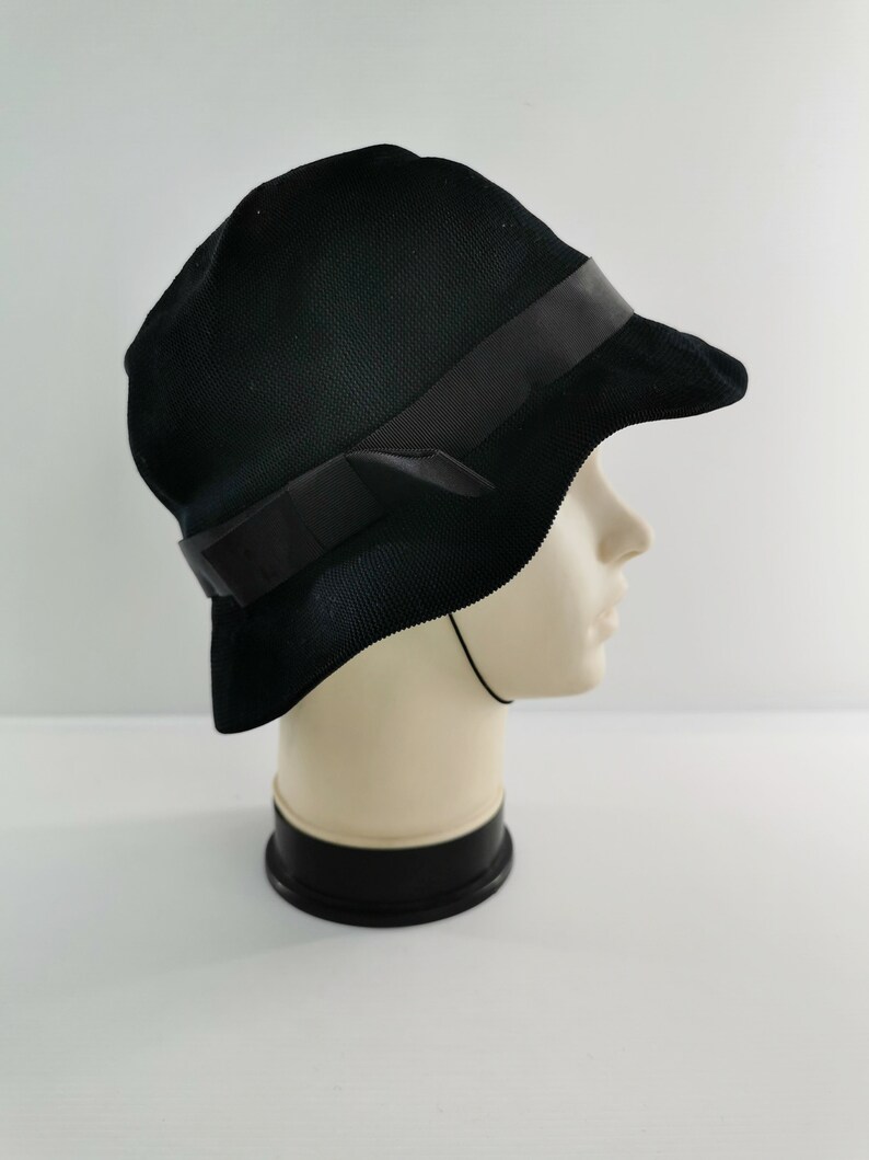 May include: A black cloche hat with a dark ribbon band and bow detail. The hat is made of a textured fabric and has a slightly curved brim. The hat is displayed on a mannequin head.