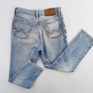 Puede incluir: Vaqueros de mezclilla azul claro con un lavado descolorido. Los vaqueros presentan un estilo cl&aacute;sico de cinco bolsillos, un parche de cuero y un dise&ntilde;o distintivo en los bolsillos traseros. Los vaqueros est&aacute;n colocados sobre una superficie blanca.