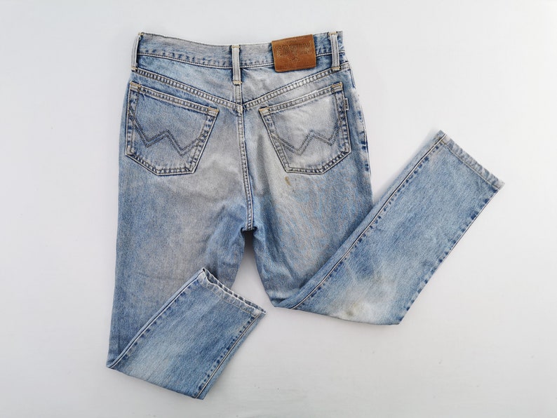 Puede incluir: Vaqueros de mezclilla azul claro con un lavado descolorido. Los vaqueros presentan un estilo cl&aacute;sico de cinco bolsillos, un parche de cuero y un dise&ntilde;o distintivo en los bolsillos traseros. Los vaqueros est&aacute;n colocados sobre una superficie blanca.