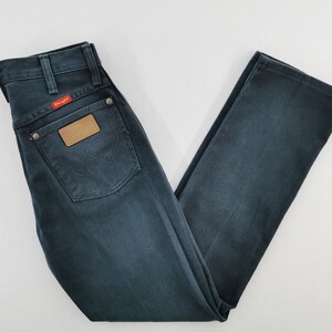 K&ouml;nnte beinhalten: Dunkelblaue Jeans aus Denim mit klassischem F&uuml;nf-Taschen-Design. Mit einem braunen Lederaufn&auml;her und einem roten "Wrangler"-Etikett. Die Jeans ist gefaltet und zeigt die Ges&auml;&szlig;tasche und das Markenlogo.