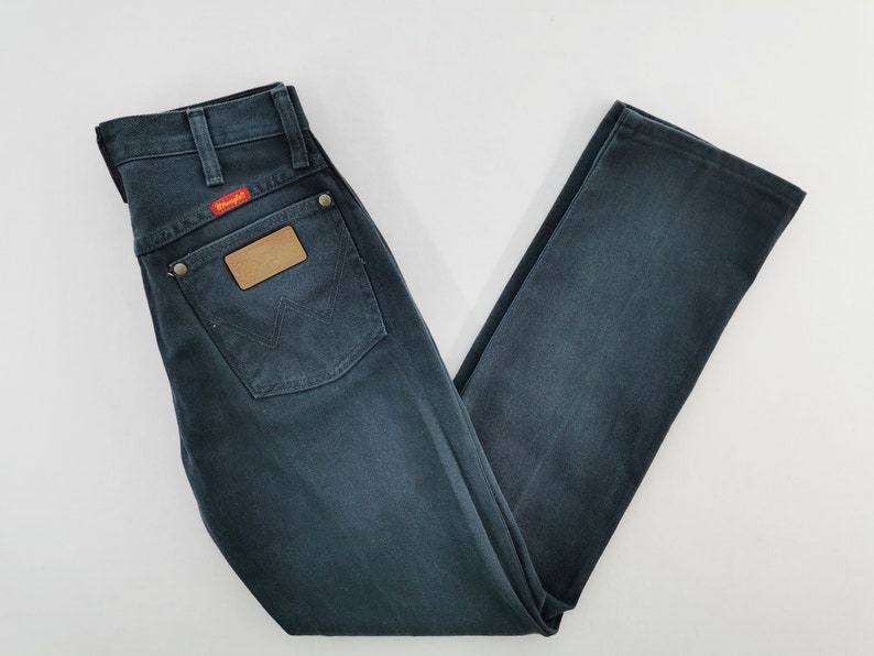 K&ouml;nnte beinhalten: Dunkelblaue Jeans aus Denim mit klassischem F&uuml;nf-Taschen-Design. Mit einem braunen Lederaufn&auml;her und einem roten "Wrangler"-Etikett. Die Jeans ist gefaltet und zeigt die Ges&auml;&szlig;tasche und das Markenlogo.