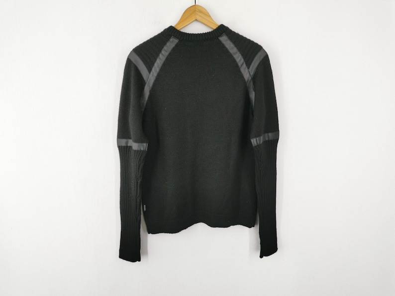 May include: Black sweater with faux leather accents on the shoulders and sleeves. The sweater is hanging on a wooden hanger against a white background. The sweater has a ribbed knit cuff and a crew neck.