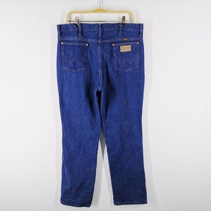 Puede incluir: Vaqueros de mezclilla azul oscuro con un corte cl&aacute;sico, con el logo de Wrangler en el bolsillo trasero y un parche de cuero. Los vaqueros cuelgan de una percha dorada sobre un fondo blanco.