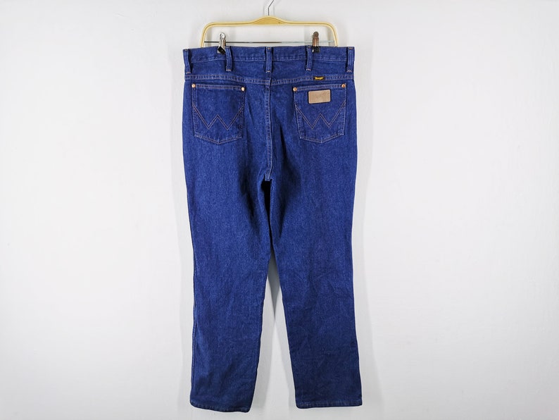Puede incluir: Vaqueros de mezclilla azul oscuro con un corte cl&aacute;sico, con el logo de Wrangler en el bolsillo trasero y un parche de cuero. Los vaqueros cuelgan de una percha dorada sobre un fondo blanco.