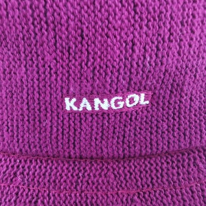 K&ouml;nnte beinhalten: Nahaufnahme einer magenta-farbenen Kangol-M&uuml;tze. Die M&uuml;tze besteht aus einem Strickstoff mit strukturierter Optik. Der Markenname "KANGOL" ist wei&szlig; auf einem rechteckigen Aufn&auml;her gestickt.