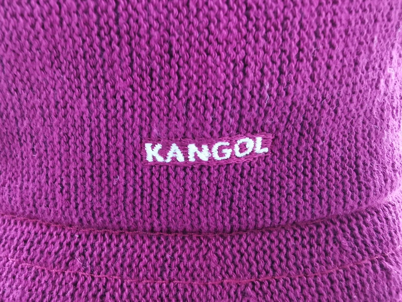 K&ouml;nnte beinhalten: Nahaufnahme einer magenta-farbenen Kangol-M&uuml;tze. Die M&uuml;tze besteht aus einem Strickstoff mit strukturierter Optik. Der Markenname "KANGOL" ist wei&szlig; auf einem rechteckigen Aufn&auml;her gestickt.