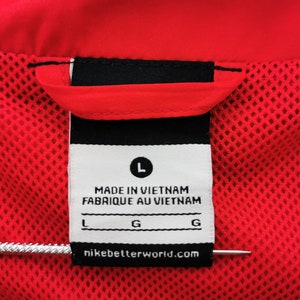May include: Close-up of a red garment tag with the text "MADE IN VIETNAM FABRIQUE AU VIETNAM". The tag includes the size "L" and the website "nikebetterworld.com". The garment has a red mesh lining.
