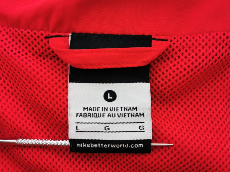 May include: Close-up of a red garment tag with the text "MADE IN VIETNAM FABRIQUE AU VIETNAM". The tag includes the size "L" and the website "nikebetterworld.com". The garment has a red mesh lining.