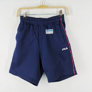 Puede incluir: Pantalones cortos deportivos azul marino con una raya roja en el lateral y el nombre de la marca "FILA" en blanco. Los pantalones cortos tienen una cintura el&aacute;stica y un peque&ntilde;o parche con texto. Los pantalones cortos est&aacute;n colgados en una percha de madera.