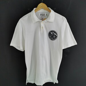 Puede incluir: Polo blanco con mangas cortas, cuello y tapeta de tres botones. La camisa presenta un gr&aacute;fico circular en blanco y negro en el pecho izquierdo. La marca "AVIREX LTD" es visible en la etiqueta.
