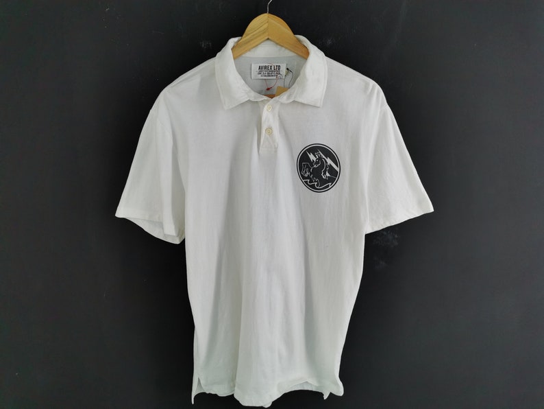 Puede incluir: Polo blanco con mangas cortas, cuello y tapeta de tres botones. La camisa presenta un gr&aacute;fico circular en blanco y negro en el pecho izquierdo. La marca "AVIREX LTD" es visible en la etiqueta.