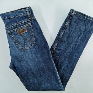 May include: Dark blue denim jeans with a classic five-pocket design. The jeans feature a brown leather patch on the back pocket with the Wrangler logo and a stitched design. The jeans are laid flat on a white surface.