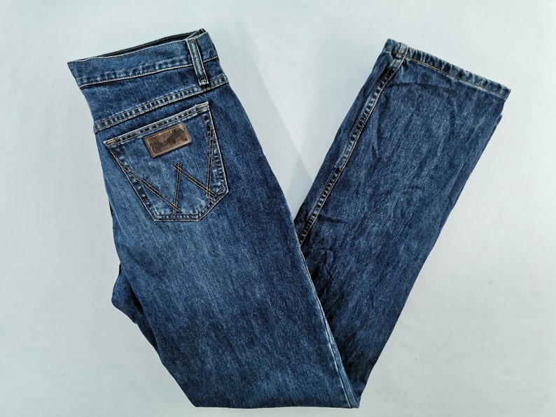 May include: Dark blue denim jeans with a classic five-pocket design. The jeans feature a brown leather patch on the back pocket with the Wrangler logo and a stitched design. The jeans are laid flat on a white surface.