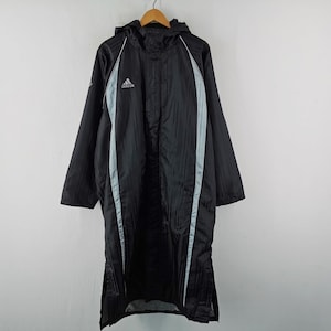 Puede incluir: Chubasquero Adidas negro con capucha y rayas azul claro. El abrigo tiene capucha y el logo de Adidas en el pecho. Cuelga de una percha de madera. Mide aproximadamente 150 cm de largo.