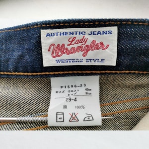Op de afbeelding: Close-up van een blauwe denim jeans met zichtbare stiksels en labels. De labels luiden "AUTHENTIC JEANS Lady Wrangler WESTERN STYLE" en bevatten maat- en wasvoorschriften. De jeans is gemaakt van 100% katoen.