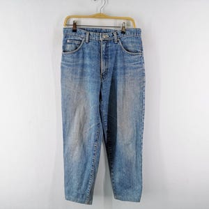 Op de afbeelding: Lichtblauwe spijkerbroek hangend aan een houten hanger tegen een witte muur. De jeans heeft een klassiek ontwerp met vijf zakken en een vervaagde wassing. De jeans is ongeveer 76 cm lang.