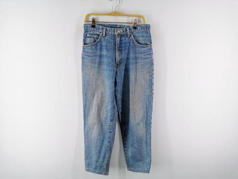 Op de afbeelding: Lichtblauwe spijkerbroek hangend aan een houten hanger tegen een witte muur. De jeans heeft een klassiek ontwerp met vijf zakken en een vervaagde wassing. De jeans is ongeveer 76 cm lang.