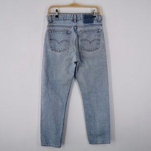 May include: Pale blue denim jeans hanging on a wooden hanger. The jeans have a classic five-pocket design, a button and zip closure, and a leather patch on the back. The back pockets feature the iconic Levi's stitching.