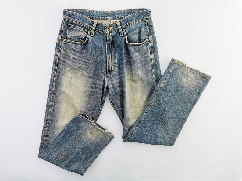 May include: Faded blue denim jeans with a classic five-pocket design. The jeans show signs of wear and tear, with distressed areas and a light wash effect. The jeans are laid flat on a white surface, showcasing their vintage style.