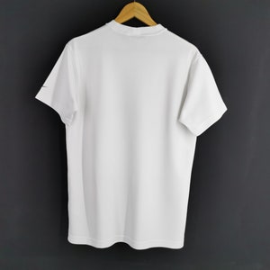 May include: A plain white t-shirt with short sleeves hangs on a wooden hanger against a dark grey background. A small black logo is visible on the left sleeve. The shirt is made of a smooth, opaque fabric.