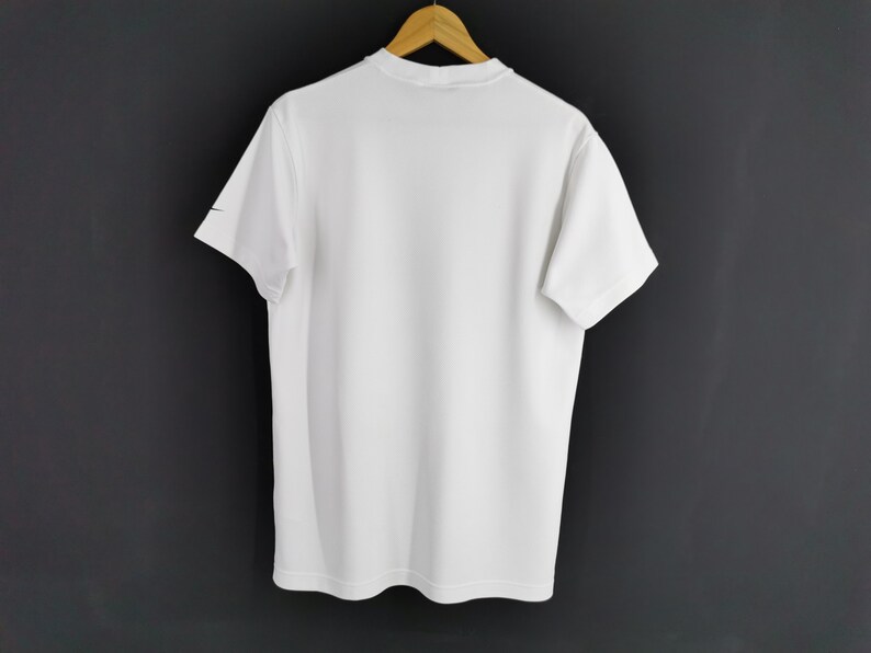 May include: A plain white t-shirt with short sleeves hangs on a wooden hanger against a dark grey background. A small black logo is visible on the left sleeve. The shirt is made of a smooth, opaque fabric.