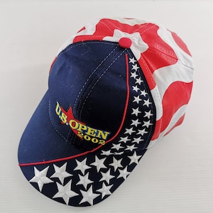Op de afbeelding: Een marineblauwe baseballcap met een rood-wit sterrenpatroon. De cap heeft de tekst "U.S. OPEN 2002" in gele en rode borduursels. Witte sterren zijn langs de rand geborduurd en er zit een rode knoop bovenop.