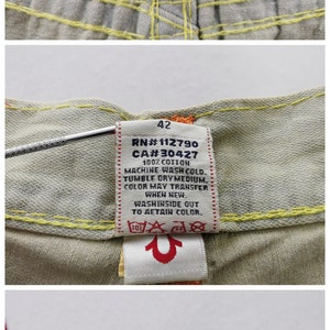 May include: Close-up of True Religion jeans labels. The labels include the brand logo, "made in USA" text, size 42, and care instructions. The jeans have yellow stitching and a small American flag patch.