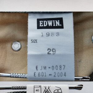May include: Close-up of light blue denim jeans with a visible label. The label reads "EDWIN 1983" and "SIZE 29". The jeans are made of 100% cotton and have a leather label. The jeans are made in Japan.