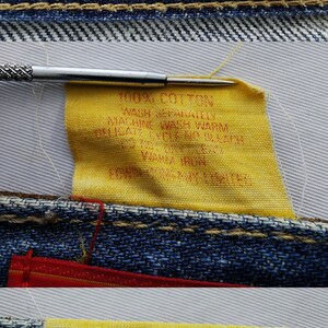 May include: Close-up of denim fabric with red and yellow labels. The red label reads "EDWIN US CLASSIC." A yellow label displays washing instructions and fabric content. A needle is positioned near the fabric.