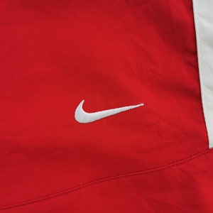 May include: Red athletic jacket with a white panel on the right side. A white Nike swoosh logo is embroidered on the left chest. The jacket appears to be made of a lightweight, possibly nylon, material.