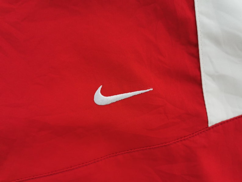 May include: Red athletic jacket with a white panel on the right side. A white Nike swoosh logo is embroidered on the left chest. The jacket appears to be made of a lightweight, possibly nylon, material.