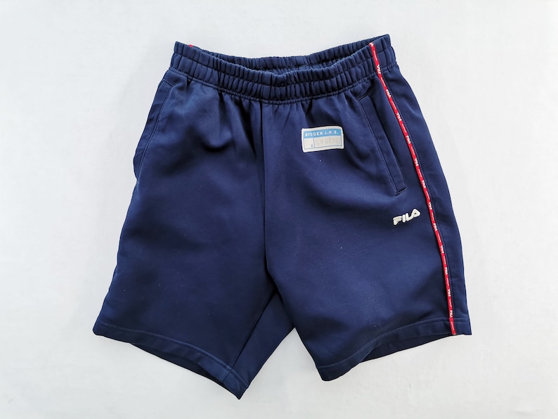 Puede incluir: Pantalones cortos deportivos azul marino con cintura el&aacute;stica y logotipo "FILA" blanco. Los pantalones cortos tienen una raya roja y blanca en el lateral y un peque&ntilde;o parche con texto. Adecuados para actividades deportivas.