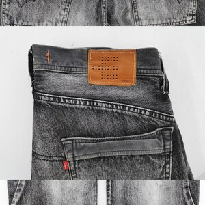 May include: Close-up of gray denim jeans with a faded wash. Features include back pockets, a leather patch with the brand name, and orange stitching details. The jeans are folded, showcasing the back and part of the legs.