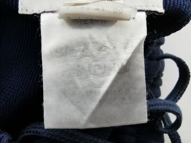 Puede incluir: Primer plano de una etiqueta de tela blanca con texto descolorido, adherida a una prenda azul marino. La etiqueta es rectangular con un borde doblado. Se ve un cord&oacute;n azul marino.