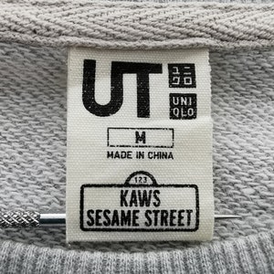 K&ouml;nnte beinhalten: Nahaufnahme eines grauen Sweatshirt-Etiketts mit dem Text "UT" und "UNIQLO". Das Etikett enth&auml;lt auch den Text "M", "MADE IN CHINA", "123", "KAWS" und "SESAME STREET".