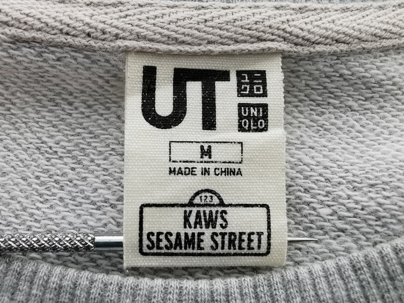 K&ouml;nnte beinhalten: Nahaufnahme eines grauen Sweatshirt-Etiketts mit dem Text "UT" und "UNIQLO". Das Etikett enth&auml;lt auch den Text "M", "MADE IN CHINA", "123", "KAWS" und "SESAME STREET".