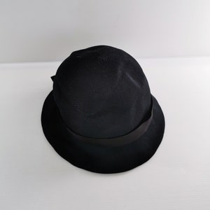 May include: A black cloche hat with a rounded crown and a small, downward-sloping brim. The hat features a black ribbon band around the crown. The hat is made of a woven material.