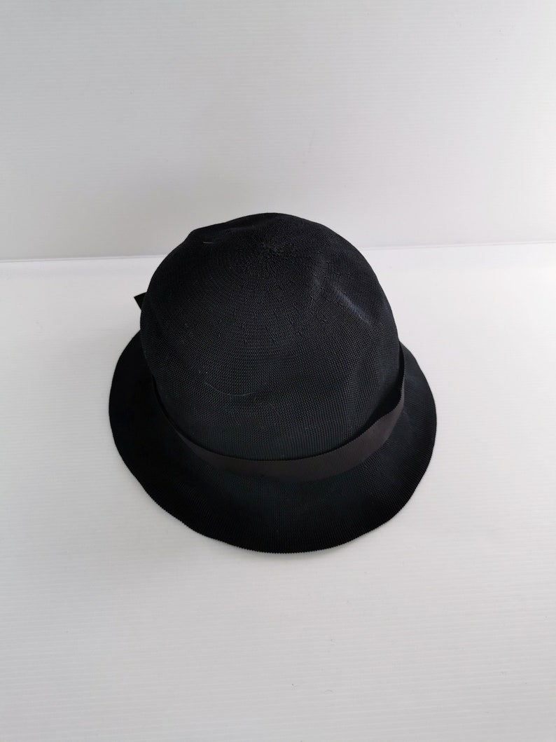 May include: A black cloche hat with a rounded crown and a small, downward-sloping brim. The hat features a black ribbon band around the crown. The hat is made of a woven material.