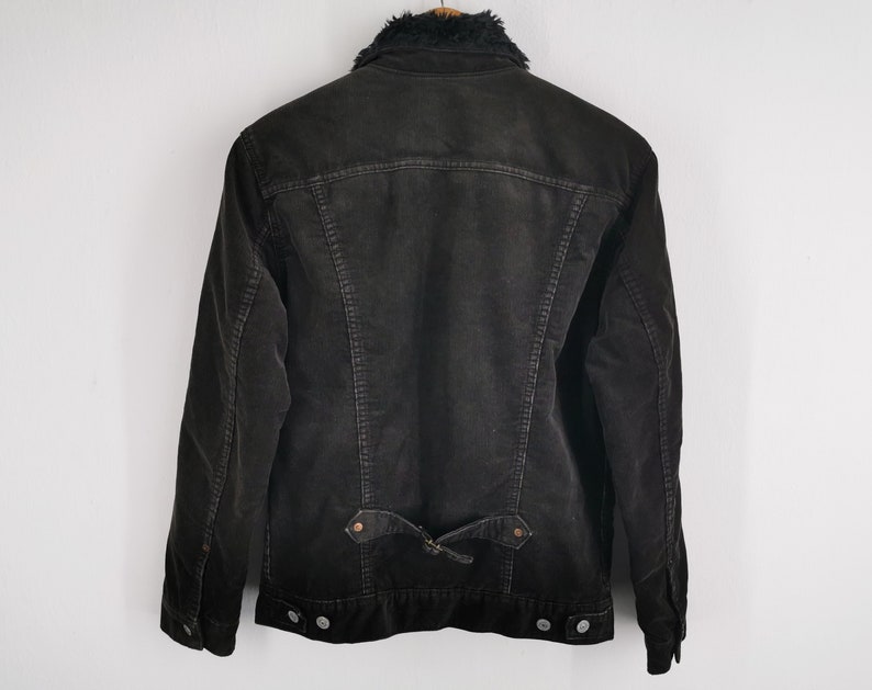 Puede incluir: Chaqueta de pana gris oscuro con cuello de piel sint&eacute;tica negra. La chaqueta tiene cierre de botones, dos bolsillos delanteros y un detalle de cintur&oacute;n en la espalda. La chaqueta se muestra sobre un fondo blanco.