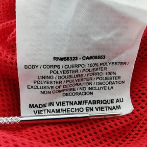 May include: Close-up of a white tag with black text, including RN#56323-CA#05553, and fabric composition details. The tag is against a red mesh fabric. The tag reads "MADE IN VIETNAM".