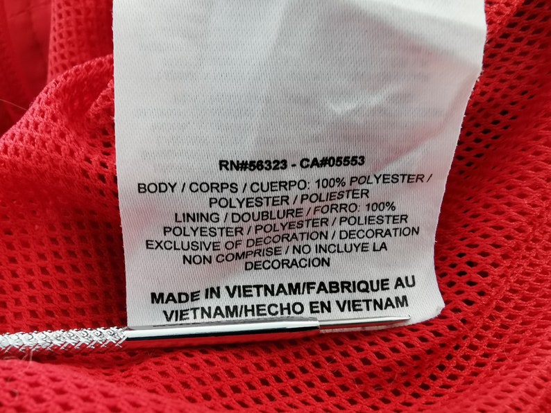 May include: Close-up of a white tag with black text, including RN#56323-CA#05553, and fabric composition details. The tag is against a red mesh fabric. The tag reads "MADE IN VIETNAM".