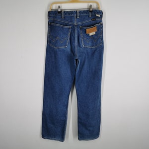 May include: Blue denim jeans with a straight leg cut, hanging on a wooden hanger. The jeans have two back pockets with decorative stitching and a leather patch. The jeans are a medium wash and appear to be vintage.