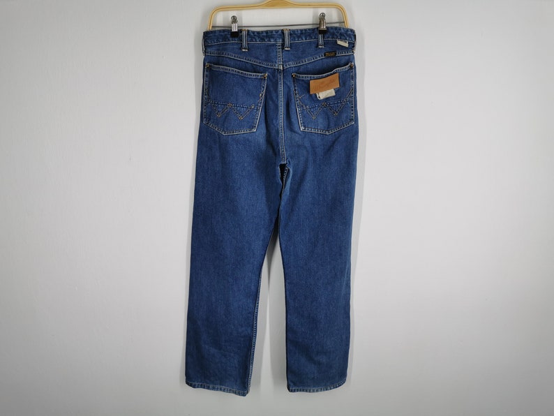 May include: Blue denim jeans with a straight leg cut, hanging on a wooden hanger. The jeans have two back pockets with decorative stitching and a leather patch. The jeans are a medium wash and appear to be vintage.