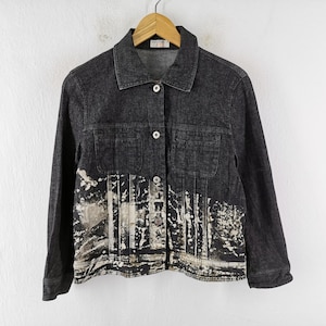 Puede incluir: Chaqueta vaquera gris oscuro con cierre de botones, dos bolsillos en el pecho y un diseño abstracto único en la parte inferior delantera. La chaqueta está colgada en una percha de madera contra una pared blanca.