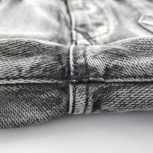 May include: Close-up of the waistband and fly of a pair of faded black denim jeans. The fabric has a textured appearance with visible stitching in black thread. The jeans are slightly wrinkled, showing wear.