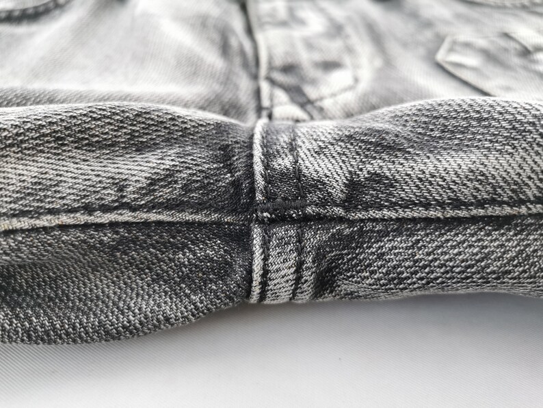May include: Close-up of the waistband and fly of a pair of faded black denim jeans. The fabric has a textured appearance with visible stitching in black thread. The jeans are slightly wrinkled, showing wear.